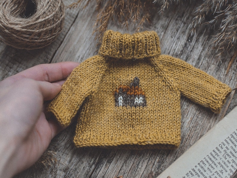 May include: A small, mustard yellow knitted jumper with a brown and white embroidered design on the front. The jumper has a turtleneck.