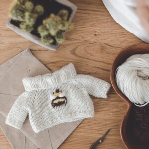 May include: A white knitted jumper with a bee embroidered on the front. The jumper is lying on a brown envelope with a pair of antique scissors nearby.