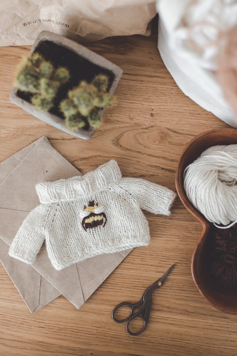 May include: A white knitted jumper with a bee embroidered on the front. The jumper is lying on a brown envelope with a pair of antique scissors nearby.