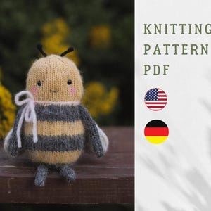 Peut inclure: Une poupée abeille tricotée avec un visage souriant, des joues roses et des antennes noires. L'abeille a une tête jaune, un corps rayé gris et jaune, et de petites ailes et pieds gris. Le texte "KNITTING PATTERN PDF" est affiché.