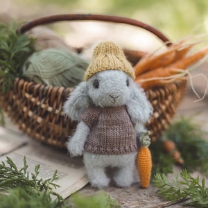 Puede incluir: Un conejito de punto gris que lleva un gorro de punto amarillo y un jersey de punto marr&oacute;n sostiene una zanahoria de ganchillo. El conejito est&aacute; sentado sobre una superficie de madera con vegetaci&oacute;n verde en el fondo.
