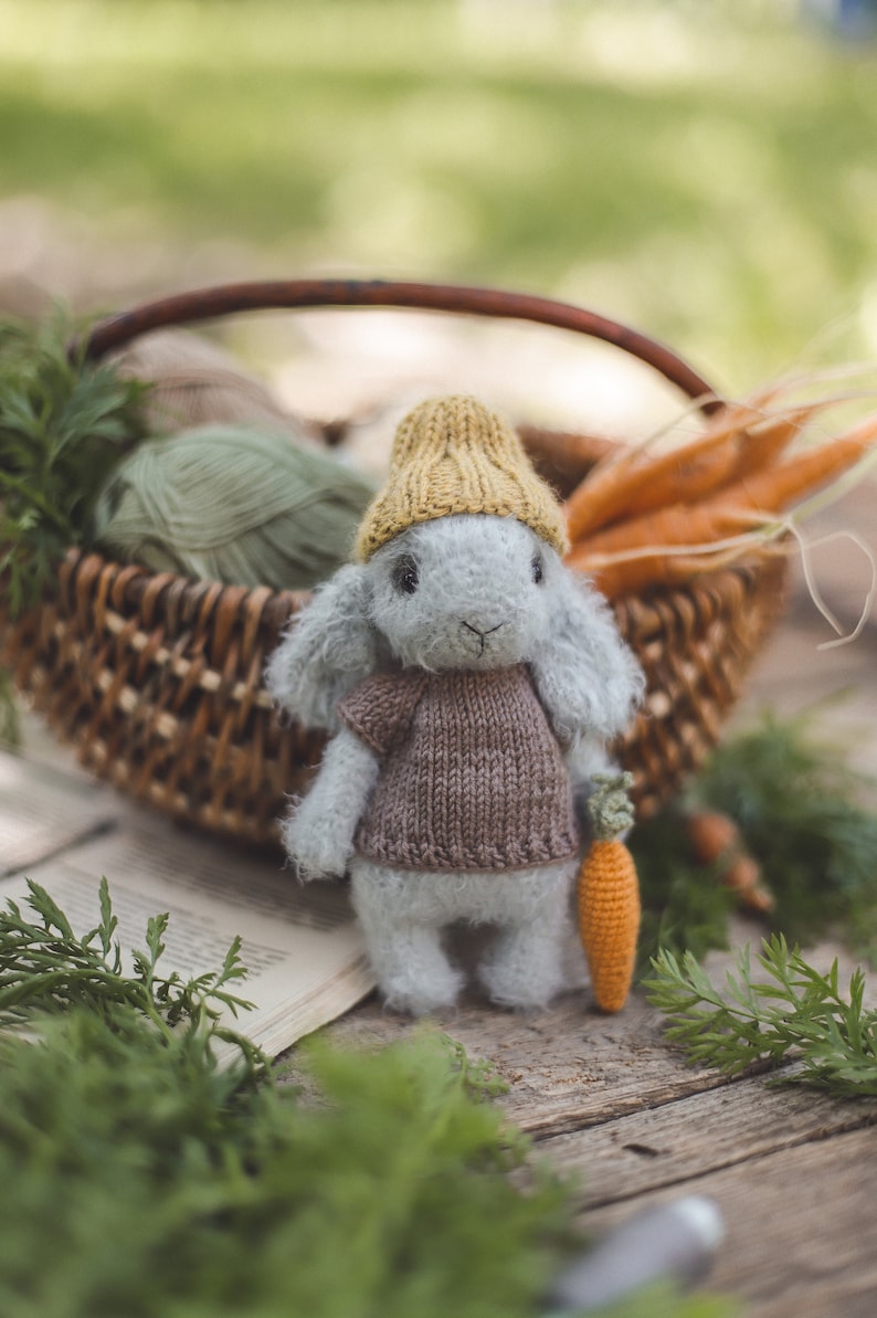 Puede incluir: Un conejito de punto gris que lleva un gorro de punto amarillo y un jersey de punto marr&oacute;n sostiene una zanahoria de ganchillo. El conejito est&aacute; sentado sobre una superficie de madera con vegetaci&oacute;n verde en el fondo.