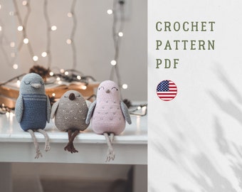 May include: Three handmade crochet bird dolls in blue, brown, and pink, with closed eyes and dangling legs, sit on a white surface. The image includes text that reads "CROCHET PATTERN PDF" and a US flag. The birds are a charming, whimsical craft.