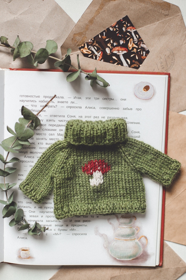 May include: A green knitted jumper with a white mushroom design. The jumper is lying on a book with text in Russian.