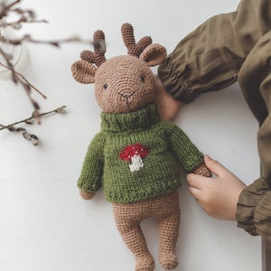 May include: A brown crocheted deer wearing a green sweater with a red and white mushroom design. The deer is lying on a white surface.