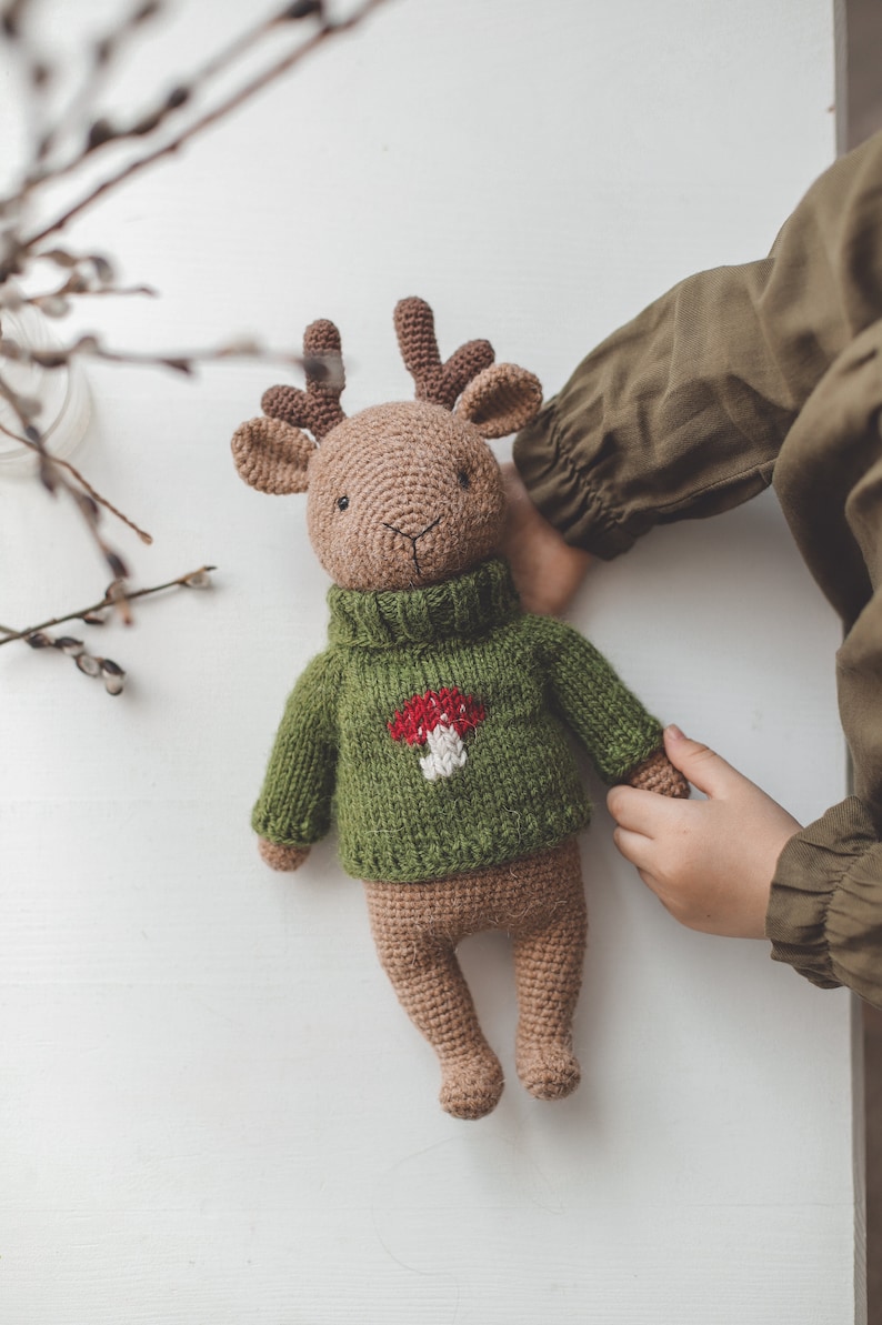 May include: A brown crocheted deer wearing a green sweater with a red and white mushroom design. The deer is lying on a white surface.