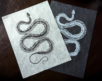 May include: Two prints featuring snake skeleton illustrations. One print has a black snake skeleton on a light gray background. The other print has a white snake skeleton on a dark gray background. The snake skeletons are detailed.