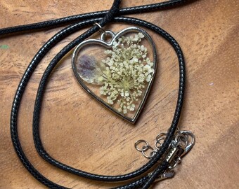 May include: A heart-shaped pendant necklace with dried flowers encased in clear resin. The pendant is framed in silver metal and hangs from a black cord necklace. The necklace is displayed on a wooden surface.