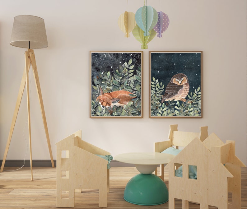 May include: Two framed illustrations of a fox and an owl sleeping in a forest setting with a starry night sky. The illustrations are hung on a white wall with a wooden floor and a wooden lamp.