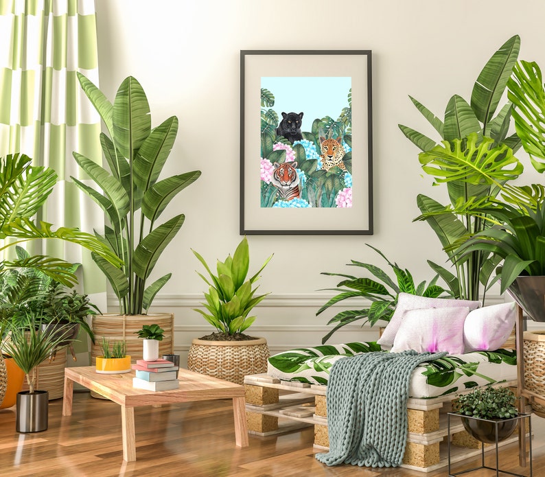 May include: A living room interior with a framed print of three tigers in a jungle setting. The print is hanging on a white wall above a wooden pallet couch with a green throw blanket. There are several potted plants in the room, including a large green plant in a brown pot, a smaller green plant in a wicker pot, and a small potted plant on a wooden coffee table.