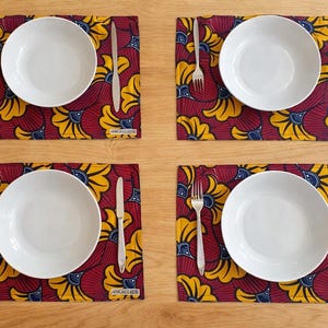 Puede incluir: Cuatro cubiertos con platos blancos, tenedores y cuchillos sobre manteles individuales estampados. Los manteles individuales presentan un fondo rojo con diseños florales amarillos y azules. La mesa está sobre una superficie de madera marrón claro.