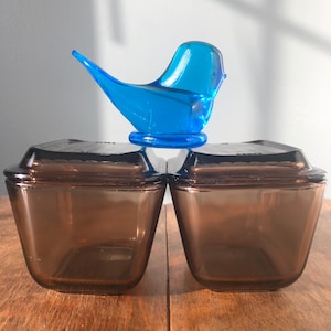 May include: Two brown glass square baking dishes with lids, stacked on top of each other. A blue glass bird figurine sits on top of the dishes.