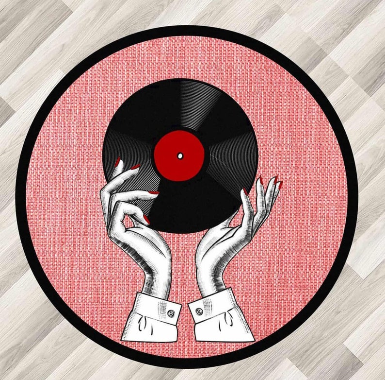 Puede incluir: Una alfombra redonda rosa y negra con un disco de vinilo negro sostenido por dos manos con esmalte de u&ntilde;as rojo. El disco tiene una etiqueta central roja.