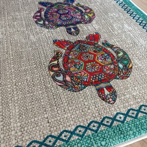 Pu&ograve; includere: Un tappeto beige con un bordo turchese e un motivo ripetitivo di tartarughe marine colorate e dettagliate. Le tartarughe sono disposte in fila, con la tartaruga pi&ugrave; grande in fondo al tappeto.