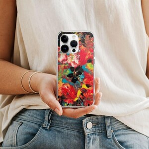 May include: A clear smartphone case featuring a vibrant, abstract paint pour design with swirling patterns of red, yellow, blue, and black. The case has a precise cutout for the triple-lens camera system and is shown held against a white top.