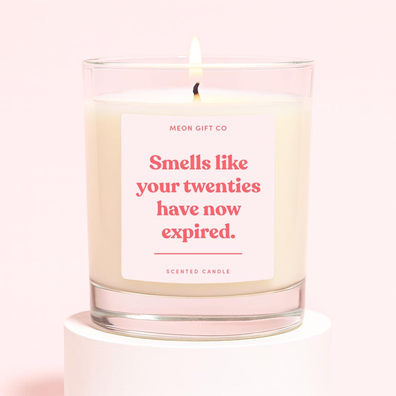 May include: A lit scented candle in a clear glass jar. The candle has a white wax and a pink label with the text "Smells like your twenties have now expired." The label also includes the text "MEON GIFT CO" and "SCENTED CANDLE."