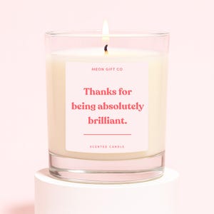 Op de afbeelding: Een brandende geurkaars in een heldere glazen pot. De kaars heeft een witte was en een roze label met de tekst "Thanks for being absolutely brilliant." De woorden "Scented Candle" staan onderaan het label.