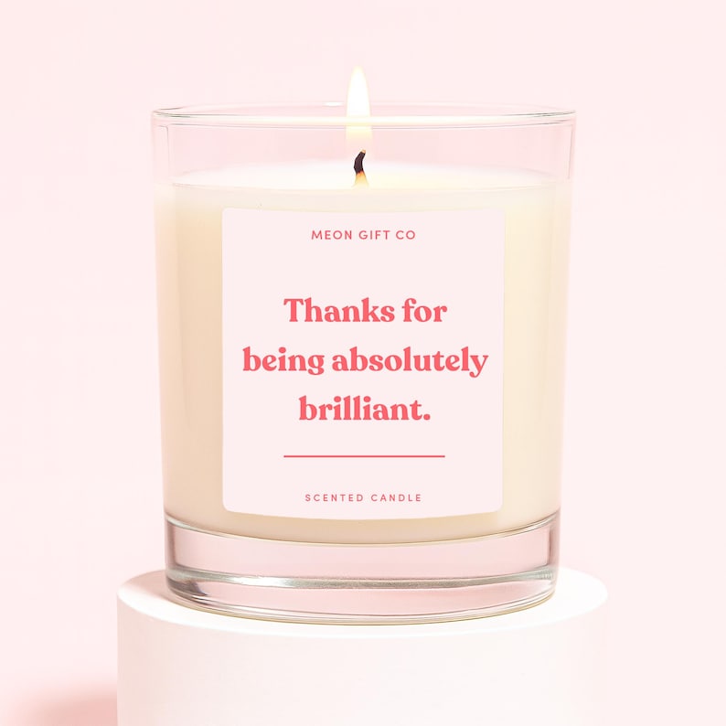 Op de afbeelding: Een brandende geurkaars in een heldere glazen pot. De kaars heeft een witte was en een roze label met de tekst "Thanks for being absolutely brilliant." De woorden "Scented Candle" staan onderaan het label.