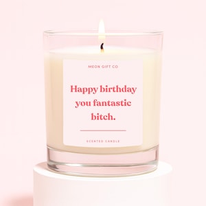 May include: A lit scented candle in a clear glass jar. The candle has a white wax and a pink label with the text "Happy birthday you fantastic bitch." The words "MEON GIFT CO" and "SCENTED CANDLE" are also printed on the label.