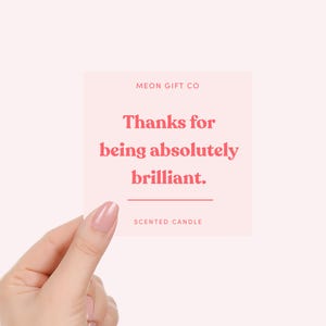 Op de afbeelding: Een vierkante, lichtroze kaart met de tekst "Thanks for being absolutely brilliant." in koraal. De kaart bevat ook de tekst "MEON GIFT CO" en "SCENTED CANDLE". De kaart wordt vastgehouden door een hand met roze nagellak.