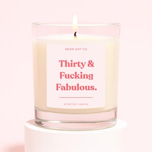 K&ouml;nnte beinhalten: Eine Duftkerze im klaren Glasgef&auml;&szlig; mit wei&szlig;em Wachs und brennendem Docht. Das Etikett zeigt den Text MEON GIFT CO, Thirty & Fucking Fabulous. sowie SCENTED CANDLE in rosa Schrift auf einem hellrosa Hintergrund auf dem Kerzenglas.