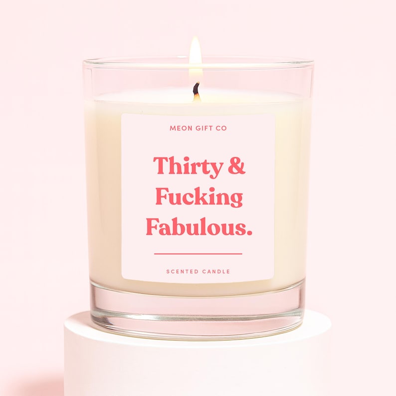 K&ouml;nnte beinhalten: Eine Duftkerze im klaren Glasgef&auml;&szlig; mit wei&szlig;em Wachs und brennendem Docht. Das Etikett zeigt den Text MEON GIFT CO, Thirty & Fucking Fabulous. sowie SCENTED CANDLE in rosa Schrift auf einem hellrosa Hintergrund auf dem Kerzenglas.
