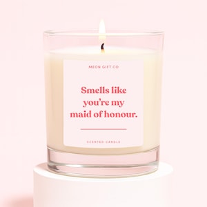 May include: A scented candle in a clear glass jar with a white label featuring the text MEON GIFT CO, Smells like you're my maid of honour, and SCENTED CANDLE in pink font. The candle has a single lit wick and white wax inside the glass container.