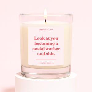 May include: A burning scented candle in a clear glass jar. The candle features a pink label with the text "Look at you becoming a social worker and shit." The brand name "MEON GIFT CO" is above the text. The words "SCENTED CANDLE" are below the text.