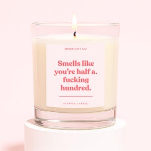 May include: A lit scented candle in a clear glass jar. The candle has a white wax and a pink label with the text "Smells like you're half a. fucking hundred." The label also includes the text "MEON GIFT CO" and "SCENTED CANDLE."