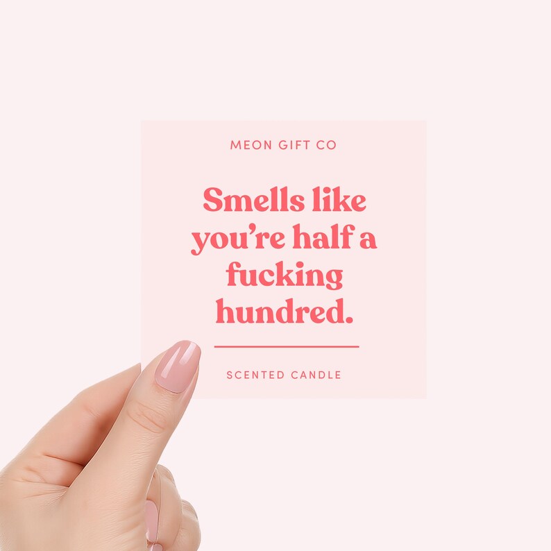 May include: A pink square card with the text "Smells like you're half a fucking hundred" in red. The card also includes the text "MEON GIFT CO" and "SCENTED CANDLE". The card is held by a hand with a pink nail polish.