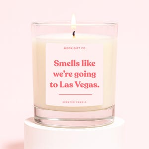 Op de afbeelding: Een brandende geurkaars in een heldere glazen pot. De kaars heeft een label met de tekst "Smells like we're going to Las Vegas." De kaars staat op een witte sokkel tegen een roze achtergrond. De tekst "Scented Candle" staat eronder.