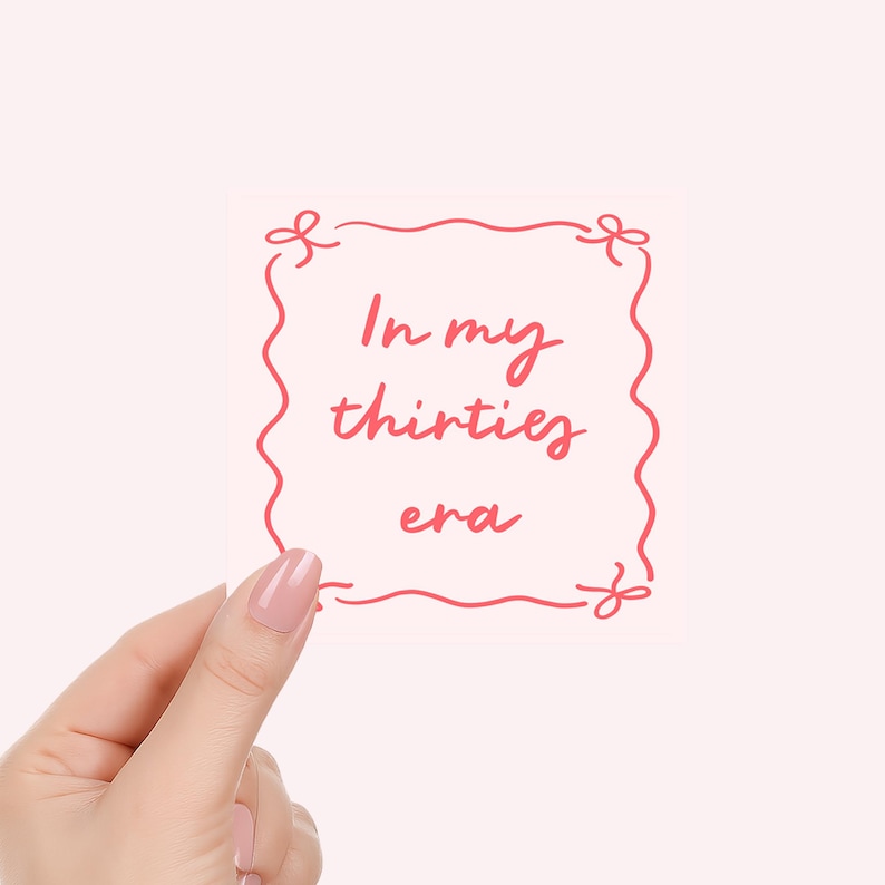 May include: A pink sticker with the text "In my thirties era" in a handwritten style, framed by a wavy red border with decorative flourishes. The sticker is held by a hand with painted nails.
