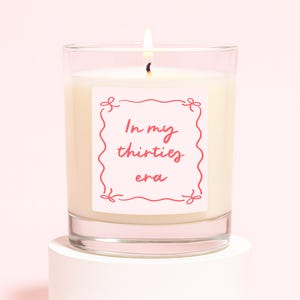 May include: A lit candle in a clear glass jar. The candle has a white wax and a label with the text "In my thirties era" in pink script. The label is framed with a pink decorative border. The candle sits on a white pedestal against a pink background.