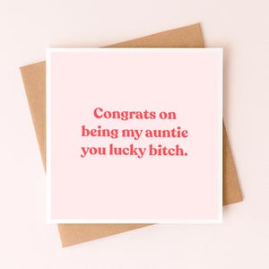 May include: A square greeting card with a light pink background and red text that reads: Congrats on being my auntie you lucky bitch. The card is placed on top of a plain brown envelope, creating a simple and direct visual presentation.