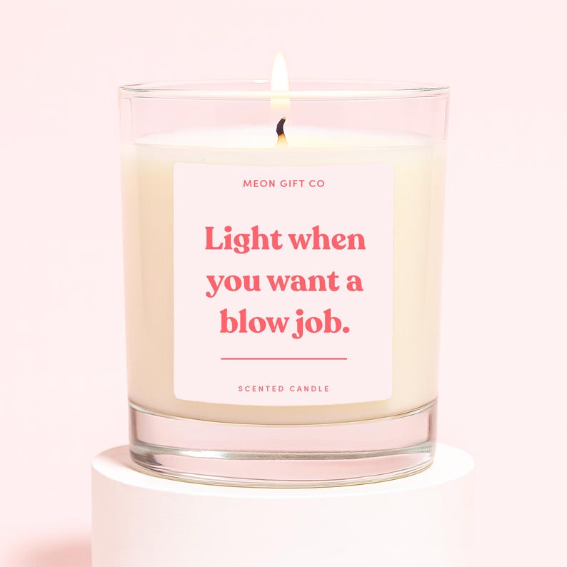 May include: A lit scented candle in a clear glass jar. The candle has a pink label with the text "Light when you want a blow job." The brand name "MEON GIFT CO" is above the text, and "SCENTED CANDLE" is below.