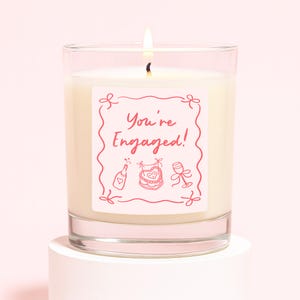 Pu&ograve; includere: Una candela accesa in un barattolo di vetro trasparente con un'etichetta rosa che recita "You're Engaged!" in una cornice decorativa. L'etichetta presenta anche illustrazioni di una bottiglia di champagne, un bavaglino e un calice. La candela &egrave; su un piedistallo bianco.