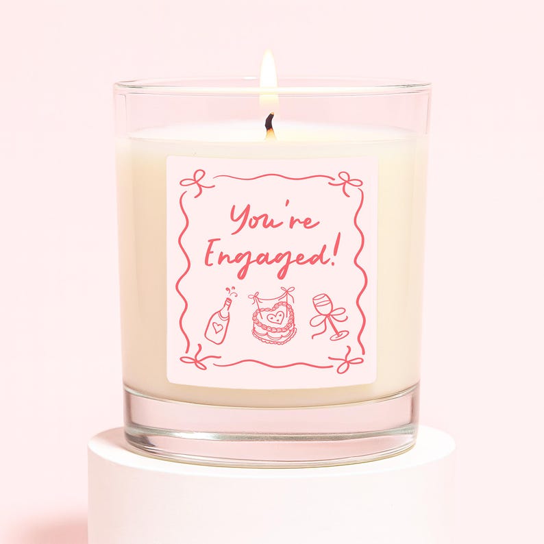 Pu&ograve; includere: Una candela accesa in un barattolo di vetro trasparente con un'etichetta rosa che recita "You're Engaged!" in una cornice decorativa. L'etichetta presenta anche illustrazioni di una bottiglia di champagne, un bavaglino e un calice. La candela &egrave; su un piedistallo bianco.
