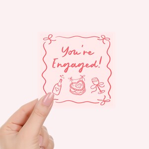 Pu&ograve; includere: Un biglietto quadrato rosa con la scritta "You're Engaged!" in rosso. Il biglietto presenta disegni a tratto di una bottiglia di champagne, una torta e un calice di vino. Il biglietto &egrave; tenuto in mano con smalto rosa.