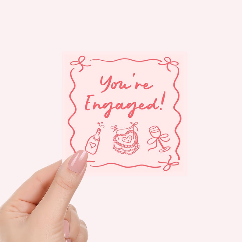 Pu&ograve; includere: Un biglietto quadrato rosa con la scritta "You're Engaged!" in rosso. Il biglietto presenta disegni a tratto di una bottiglia di champagne, una torta e un calice di vino. Il biglietto &egrave; tenuto in mano con smalto rosa.
