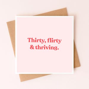 Op de afbeelding: Een vierkante wenskaart met een lichtroze achtergrond en de tekst "Thirty, flirty & thriving." gedrukt in een vet rood schreeflettertype. De kaart ligt op een eenvoudige bruine papieren envelop voor een strakke en heldere weergave.