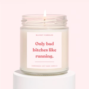 May include: A lit soy wax candle in a clear glass jar. The candle has a pink label with the text "Only bad bitches like running." The brand name "Blushy Candles" is at the top of the label, and "Handmade Soy Wax Candle" is at the bottom.
