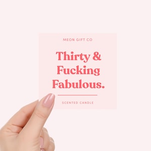 K&ouml;nnte beinhalten: Ein quadratisches, hellrosa Kerzenetikett mit dem Text MEON GIFT CO oben, gefolgt von dem Schriftzug Thirty & Fucking Fabulous. in fetter roter Schrift, einer horizontalen Linie und dem Wort SCENTED CANDLE am unteren Rand des Designs.