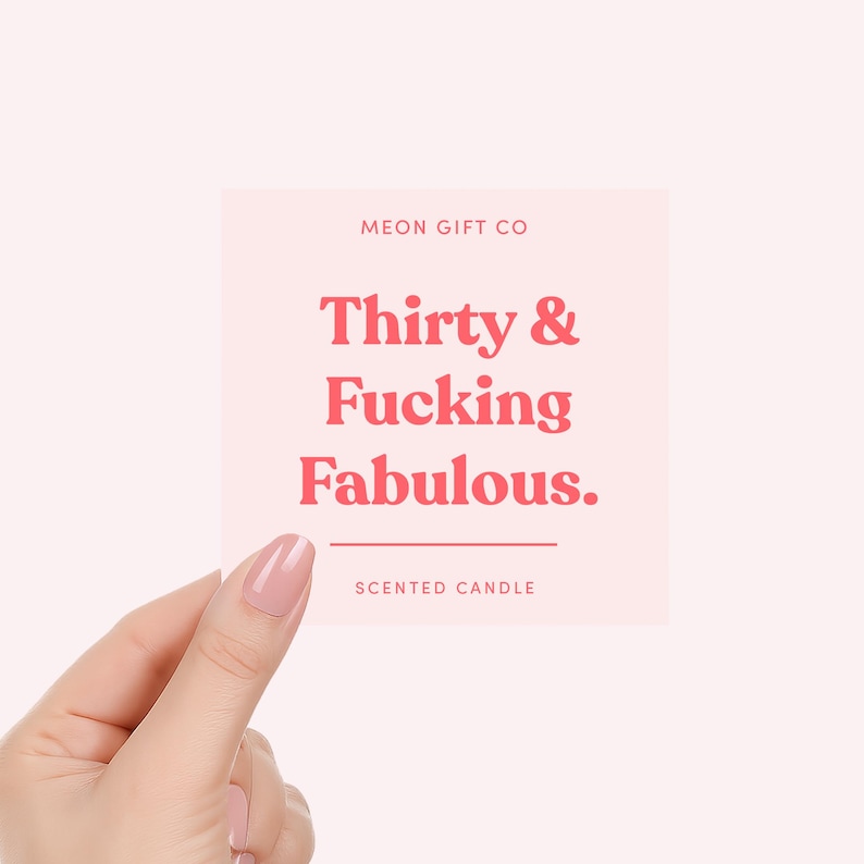 K&ouml;nnte beinhalten: Ein quadratisches, hellrosa Kerzenetikett mit dem Text MEON GIFT CO oben, gefolgt von dem Schriftzug Thirty & Fucking Fabulous. in fetter roter Schrift, einer horizontalen Linie und dem Wort SCENTED CANDLE am unteren Rand des Designs.