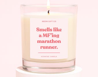 May include: A lit scented candle in a clear glass jar. The candle has a pink label with the text "Smells like a MF'ing marathon runner." The words "MEON GIFT CO" and "SCENTED CANDLE" are also printed on the label.