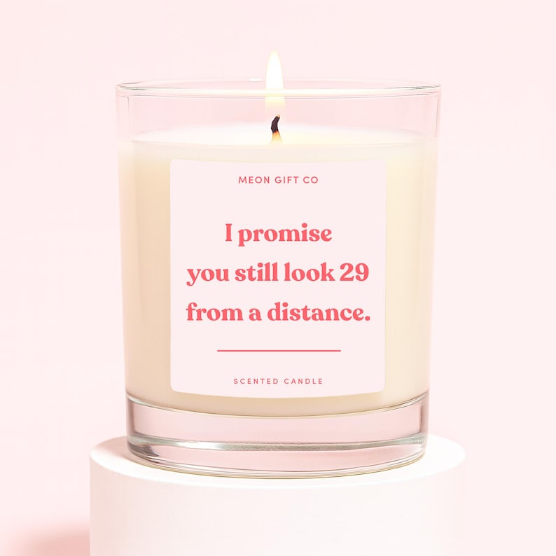 May include: A lit scented candle in a clear glass jar. The candle has a pink label with the text "I promise you still look 29 from a distance." The candle is on a white surface against a pink background.