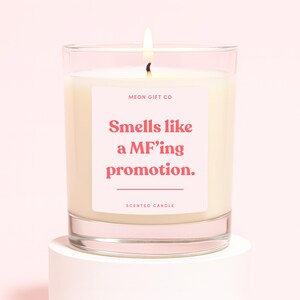 May include: A lit scented candle in a clear glass jar. The candle has a white wax and a pink label with the text "Smells like a MF'ing promotion." The words "MEON GIFT CO" and "SCENTED CANDLE" are also printed on the label.