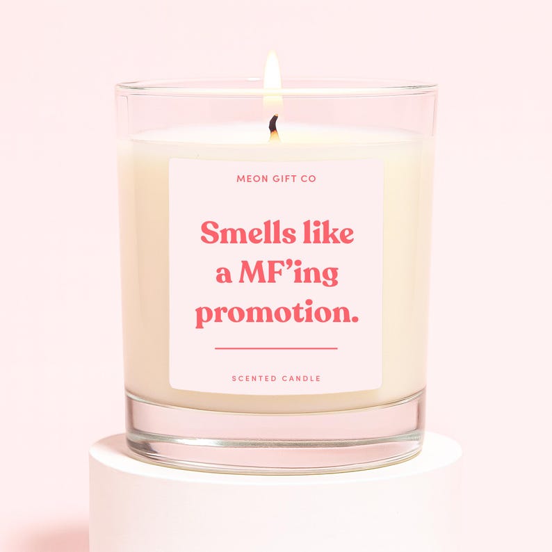 May include: A lit scented candle in a clear glass jar. The candle has a white wax and a pink label with the text "Smells like a MF'ing promotion." The words "MEON GIFT CO" and "SCENTED CANDLE" are also printed on the label.