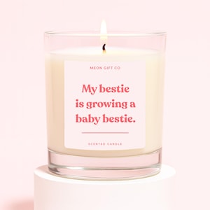 Op de afbeelding: Een brandende geurkaars in een heldere glazen pot. De kaars heeft een witte was en een roze label met de tekst "My bestie is growing a baby bestie." Het label bevat ook de tekst "Scented Candle" en het merk "MEON GIFT CO."