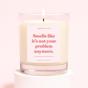 May include: A lit scented candle in a clear glass jar. The candle has a white wax and a pink label with the text "Smells like it's not your problem anymore." The label also includes the text "MEON GIFT CO" and "SCENTED CANDLE."
