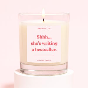May include: A lit scented candle in a clear glass jar. The candle has a white wax and a pink label with the text "Shhh... she's writing a bestseller." The label also includes the text "MEON GIFT CO" and "SCENTED CANDLE".